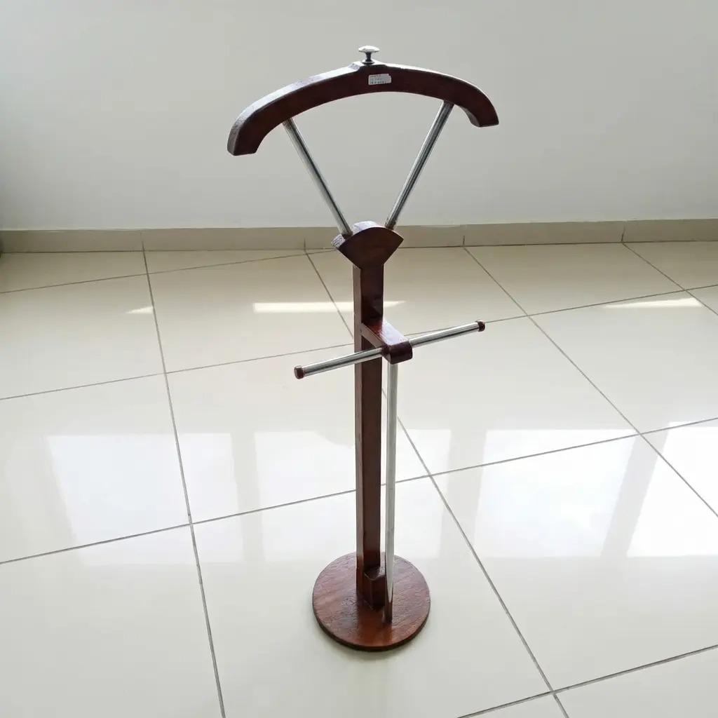 Modern freestanding wooden valet stand with a curved suit hanger, metal trouser bar, and a stable circular base in a minimalist white-tiled room.