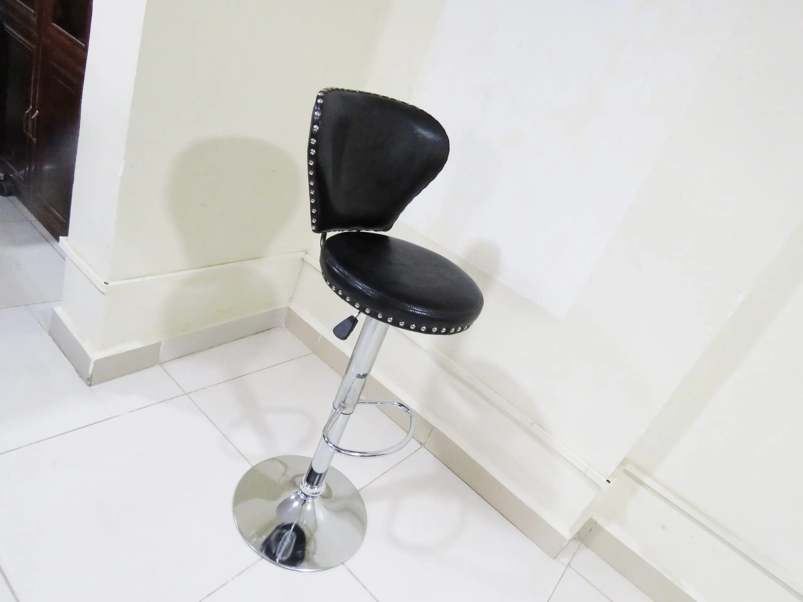 Leather swivel barstool -black featuring a curved, padded backrest and a thick round seat cushion upholstered in shiny black faux leather with silver chrome rivet stud trim, supported by an adjustable pneumatic chrome pedestal stem with a teardrop lever, an integrated semi-circular chrome footrest, and a polished chrome trumpet base.