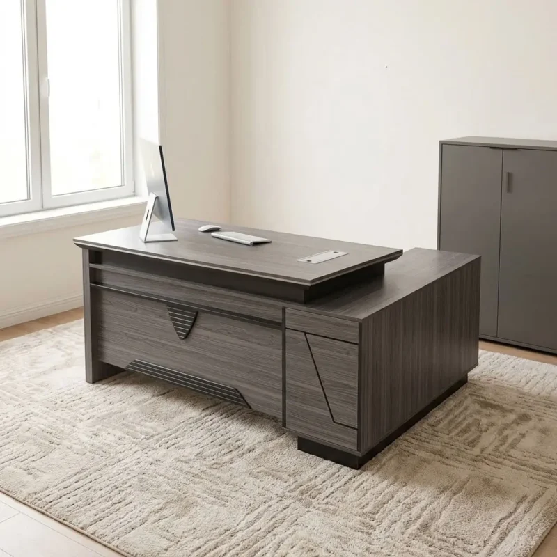 L-shaped 1800mm office table featuring a dark grey wood-grain laminate finish, dual desktop flip-top cable management ports, a front modesty panel with geometric black accent detailing, an integrated side return extension unit providing additional work surface and storage, and solid panel-style engineered wood base supports.